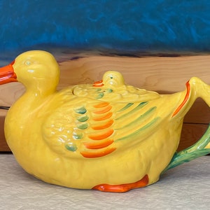 May include: A yellow ceramic teapot shaped like a duck with an orange beak and feet. The duck features green and orange painted wing details and a small duckling figure on the lid. The handle is shaped like a green reed or plant stem.