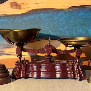 Peut inclure: Une balance à plateaux de style ancien avec une base en fonte marron foncé et deux coupelles en laiton. La balance est accompagnée de deux jeux de poids métalliques circulaires gradués, un grand jeu à gauche et un plus petit à droite.