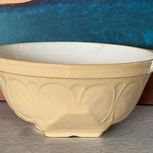 May include: A round ceramic mixing bowl with a pale yellow exterior and a smooth white interior. The outer surface features a moulded decorative pattern of repeating arches and a textured rim. The bowl has a wide, flared edge and a sturdy base.