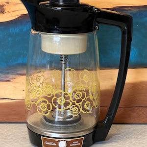 May include: A vintage Sunbeam fully automatic coffee maker. The glass carafe has a yellow floral design. The coffee maker has a black lid, handle, and base. The control dial reads "Mild" and "Strong".