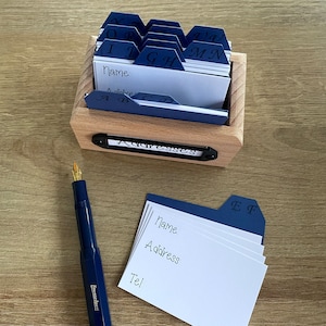 May include: A wooden box with a blue and white index card file system. The box has a label that says "Addresses". The cards are labeled with letters of the alphabet. A blue pen and a stack of blank index cards are in the foreground.