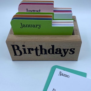 May include: A wooden box with the word "Birthdays" on the front. The box is filled with colorful cards, each with a month of the year written on it. The cards are labeled "January" and "August".