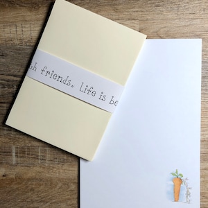 May include: A white envelope with a cream colored insert. The insert has a white ribbon with the text "With friends. Life is be" printed on it. The envelope has a white background with a drawing of two bunnies and a carrot.