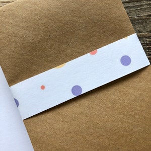May include: A brown paper envelope with a white strip of paper adhered to the bottom edge. The white paper has a repeating pattern of small purple, orange, and yellow circles.