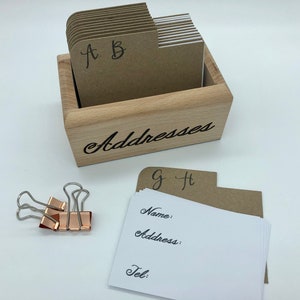 May include: A wooden box with the word "Addresses" engraved on the front. The box contains a set of index cards with alphabetical tabs, labeled A through H. The cards are blank and ready to be filled with addresses. Three binder clips are also included.