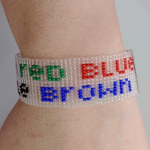 May include: A beaded bracelet with the words "RED BLUE BROWN" spelled out in colorful beads. The bracelet is white with red, green, blue, and black lettering. The bracelet is secured with a silver clasp.
