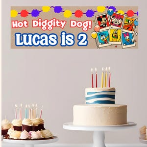 K&ouml;nnte beinhalten: Ein Geburtstagsbanner mit dem Text "Hot Diggity Dog! Lucas is 2" und Cartoon-Charakter-Bildern. Ein zweist&ouml;ckiger Kuchen mit Kerzen auf einem wei&szlig;en Kuchenst&auml;nder, sowie Cupcakes. Bunte B&auml;nder und Konfetti erg&auml;nzen die festliche Szene.