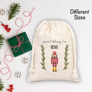 May include: A cream-coloured drawstring gift bag with the text "Special Delivery for HENRY" and a nutcracker illustration. The bag is decorated with green sprigs and a festive design. The words "Different Sizes" are also visible.