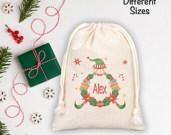 Puede incluir: Una bolsa con cordón de color crema con un diseño de elfo festivo. El elfo está rodeado por una corona con el nombre "Alex" en rojo. La bolsa está decorada con estrellas, bastones de caramelo y cajas de regalo. El texto "Different Sizes" también es visible.