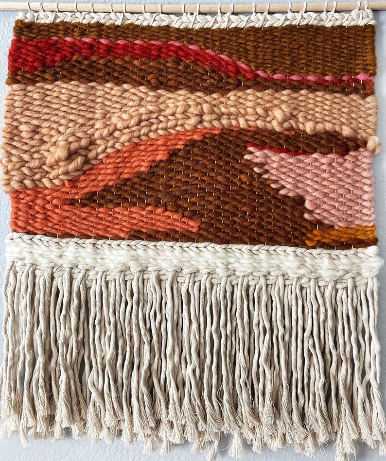 May include: A woven wall hanging with a geometric pattern in shades of brown, orange, pink, and red. The hanging has a fringe border.