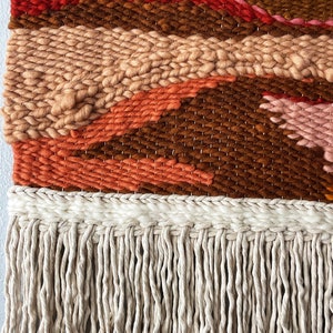 May include: A woven wall hanging with a geometric pattern in shades of brown, orange, pink, and red. The hanging has a fringe of natural fiber.