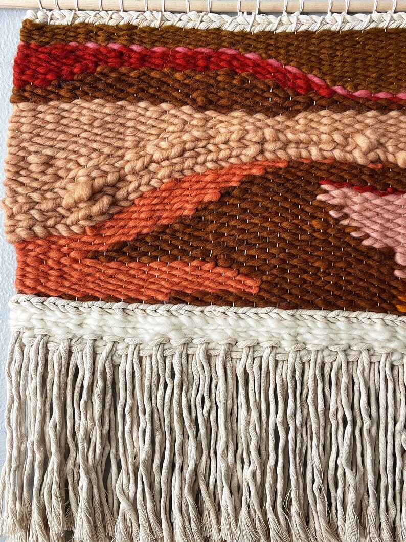 May include: A woven wall hanging with a geometric pattern in shades of brown, orange, pink, and red. The hanging has a fringe of natural fiber.