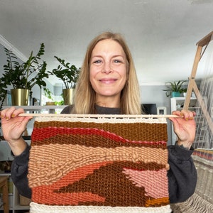 May include: A woman holds up a woven wall hanging with a colorful abstract design. The hanging is made with different shades of brown, pink, and orange yarn. The hanging has a fringe border.