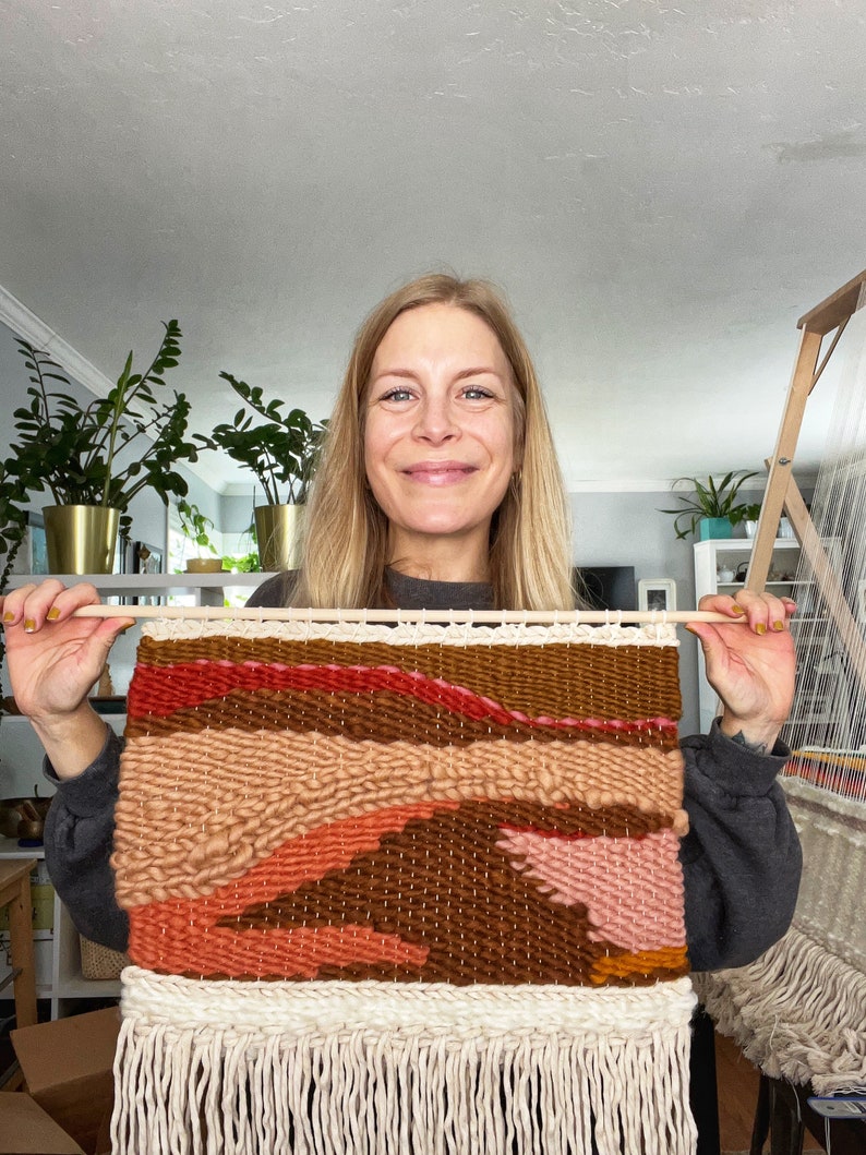 May include: A woman holds up a woven wall hanging with a colorful abstract design. The hanging is made with different shades of brown, pink, and orange yarn. The hanging has a fringe border.