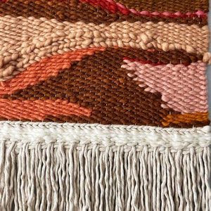 May include: A woven wall hanging with a brown, orange, pink, and red geometric pattern. The hanging has a fringe border.