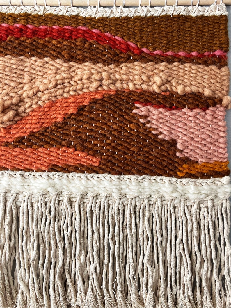 May include: A woven wall hanging with a brown, orange, pink, and red geometric pattern. The hanging has a fringe border.