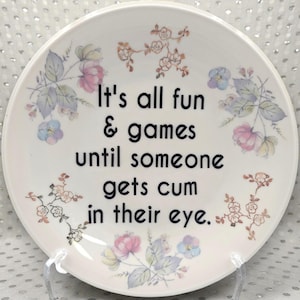 Peut inclure: Une assiette en céramique blanche ornée de motifs floraux roses, bleus et pêche. L'assiette porte l'inscription "It's all fun & games until someone gets cum in their eye" en noir.