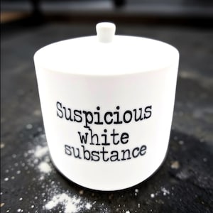 May include: A white ceramic container with a lid and the words "Suspicious white substance" printed in black. The container is cylindrical with a small knob on top.