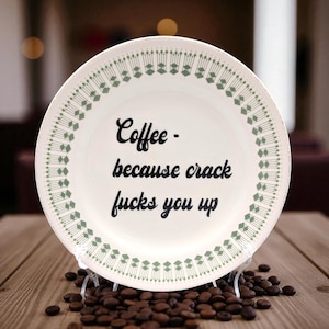 May include: A white ceramic plate with a green geometric border and the text "Coffee - because crack fucks you up" in black script. The plate rests on a clear stand, surrounded by coffee beans, on a wooden surface.