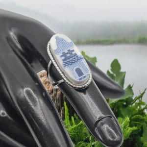 May include: An oval ring featuring a white ceramic inlay with a blue transferware-style pagoda pattern. The ceramic piece is set within a silver-toned metal bezel with a beaded edge, mounted on a thin, plain silver-toned metal band.