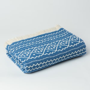 May include: A blue and white patterned throw blanket with a fringe border. The blanket is folded and lying on a white surface.