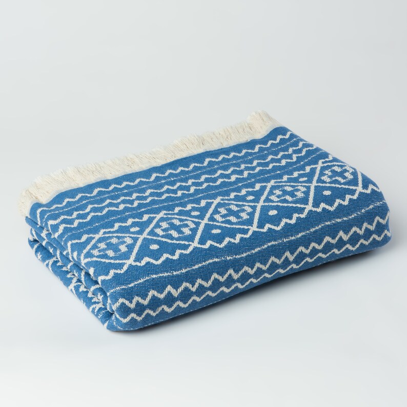 May include: A blue and white patterned throw blanket with a fringe border. The blanket is folded and lying on a white surface.