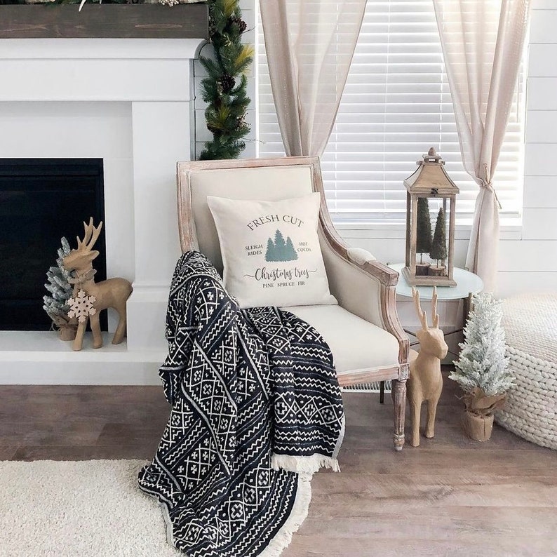 May include: A white armchair with a black and white throw blanket and a pillow with the text "Fresh Cut Sleigh Rides Hot Cocoa Christmas Trees Pine Spruce Fir" in a farmhouse style living room.