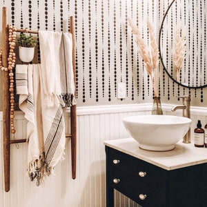 May include: A bathroom scene featuring a wooden towel ladder with cream-colored towels and a small potted plant. The wall has a black dot pattern. A white sink sits atop a black vanity with silver knobs. A round mirror and decorative vase are also present.