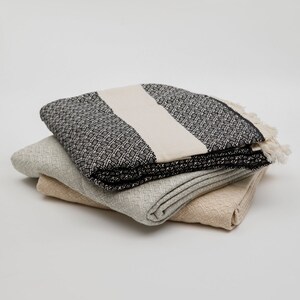 May include: A stack of three folded woven blankets. The top blanket features a black and white geometric pattern with a solid cream-coloured stripe and fringed edges. The middle blanket is light grey, and the bottom blanket is a solid tan colour.