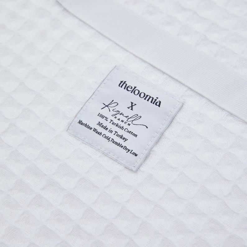 May include: White label with black text that reads "theloomia" and "Rignell Ranch" with the text "X" between the two names. The label also includes the text "100% Turkish Cotton", "Made in Turkey", and "Machine Wash Cold, Tumble Dry Low".