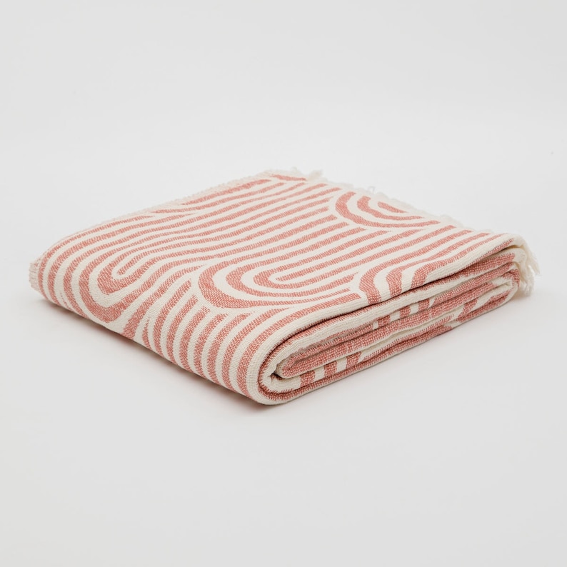 May include: A folded pink and white throw blanket with a geometric pattern of curved lines. The blanket has a fringed edge.