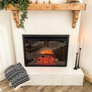 May include: A white fireplace mantel with a black electric fireplace insert. The fireplace is lit and has a realistic flame effect. The mantel is decorated with a wooden shelf and a row of wooden beads.