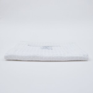 May include: A white waffle weave towel folded in thirds. The towel has a small white tag with the text "HANDMADE TOWEL" and "X" printed on it.