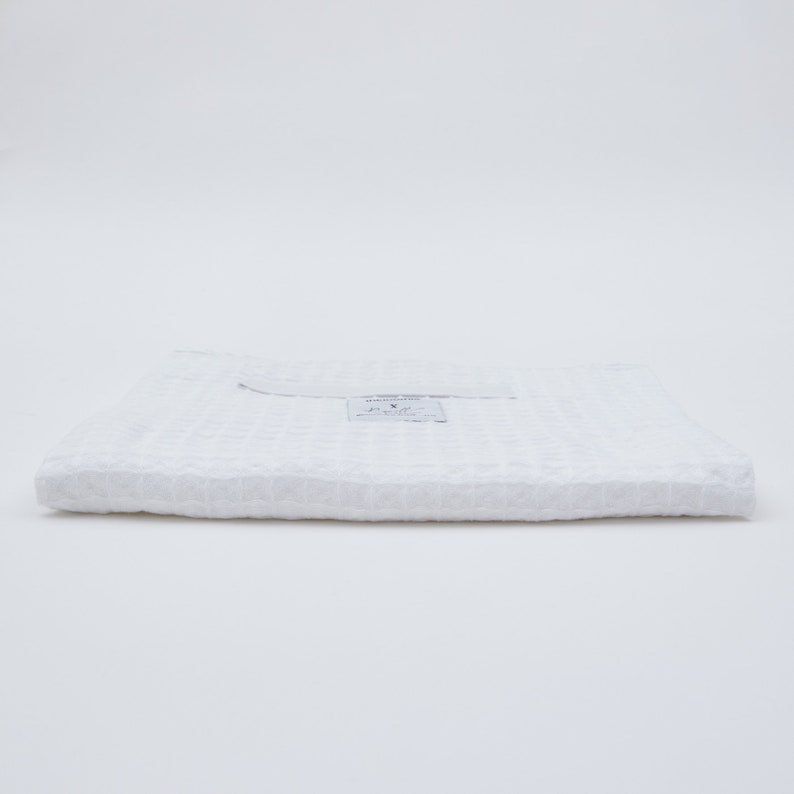May include: A white waffle weave towel folded in thirds. The towel has a small white tag with the text "HANDMADE TOWEL" and "X" printed on it.