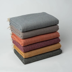 May include: A stack of seven folded blankets in various colors, including gray, tan, orange, burgundy, and gold. Each blanket has a textured appearance and a fringe detail on one edge. The blankets are neatly arranged on a white surface.