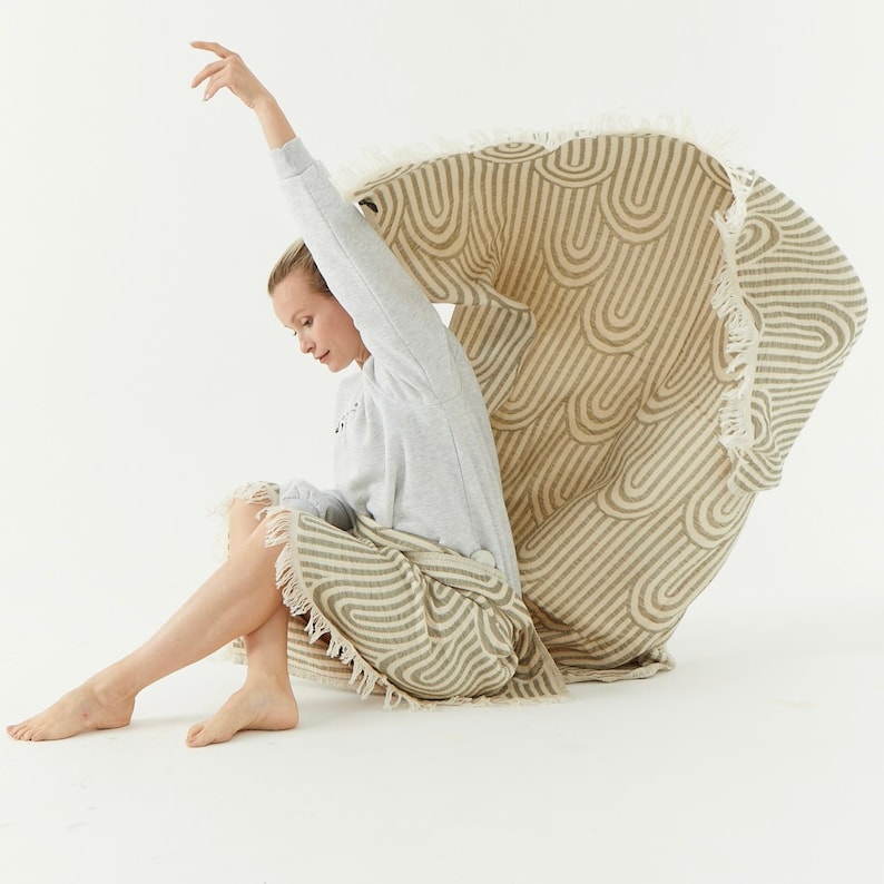 May include: A woman sits on a beige and brown striped throw blanket with fringe. The blanket has a wavy pattern.