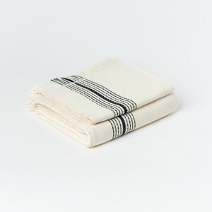 May include: Folded off-white woven textile with black stripes. The textile has a fringed edge and a textured weave. The black stripes are evenly spaced across the width of the textile. The textile is folded neatly.