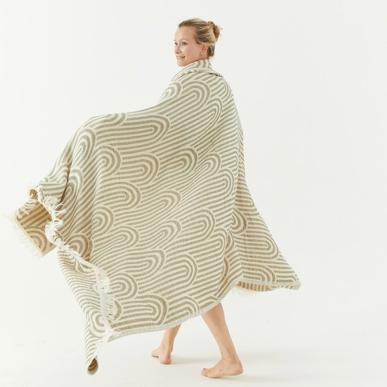 May include: A cream-colored throw blanket with a wavy, abstract pattern in a light brown color. The blanket has a fringe border and is draped over a person's shoulders.