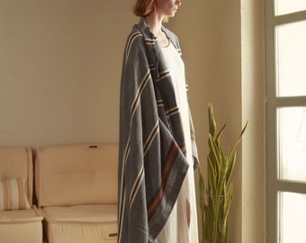 May include: A woman wearing a white dress with a blue and white striped throw blanket draped over her shoulders. The blanket has a brown stripe running down the center. The woman is standing in front of a window with a plant in a wicker basket.