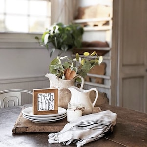 May include: A rustic wooden table setting featuring a small wooden sign that reads "fresh tulips". The scene includes white plates, a white pitcher, a glass jar with a bow, and a striped linen towel. A bouquet of greenery and a wooden heart decorates the pitcher.