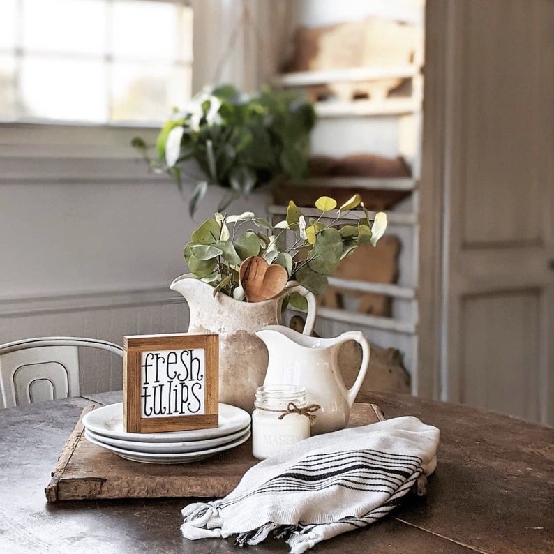 May include: A rustic wooden table setting featuring a small wooden sign that reads "fresh tulips". The scene includes white plates, a white pitcher, a glass jar with a bow, and a striped linen towel. A bouquet of greenery and a wooden heart decorates the pitcher.