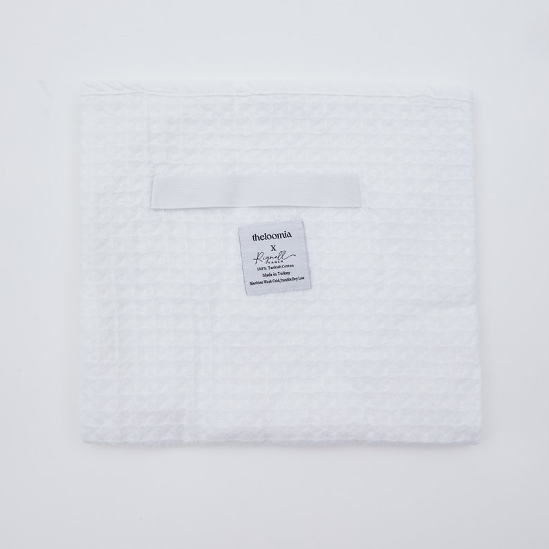 May include: White waffle weave kitchen towel with a label that reads "theloomia x Regull, 100% Turkish Cotton, Made in Turkey, Machine Wash Cold, Tumble Dry Low."