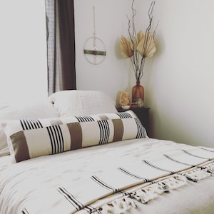 May include: A white bed with a black and brown striped bolster pillow and a white and black striped throw blanket. The bed is made with white linens and has a wooden headboard. There is a vase with dried palm leaves and a small decorative object on a side table.