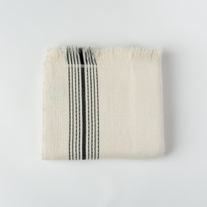 May include: Folded cream-colored woven blanket with a black vertical stripe design. The blanket has a textured surface and fringed edges. The design is simple and minimalist, suitable for home decor.