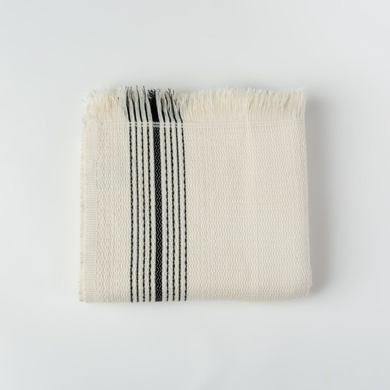 May include: Folded cream-colored woven blanket with a black vertical stripe design. The blanket has a textured surface and fringed edges. The design is simple and minimalist, suitable for home decor.
