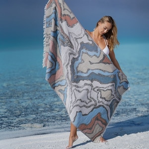 May include: A woman is holding a large beach towel with an abstract pattern in shades of blue, grey and pink. The towel has a fringed edge.