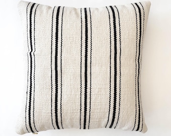 May include: Square decorative pillow with a cream-colored background and vertical black stripes. The pillow has a textured appearance and a neutral color palette, suitable for various home decor styles.