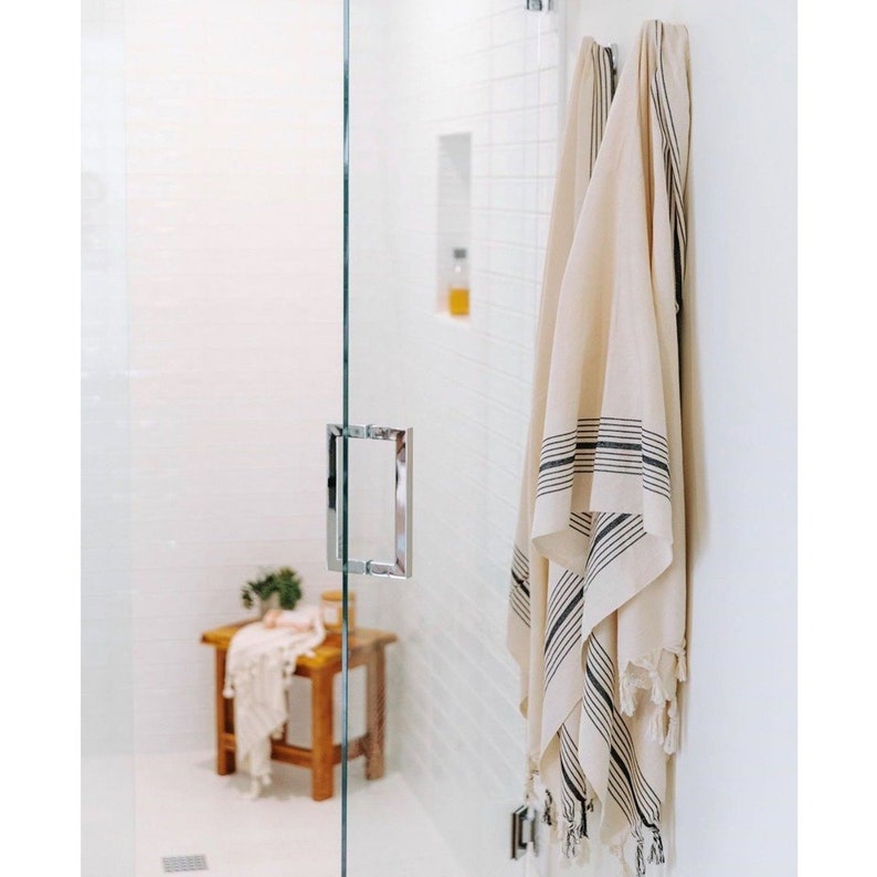 May include: A beige and black striped towel with fringe hangs on a white wall. The towel is made of a soft material and has black stripes along the edges. A wooden stool and a glass shower door are also visible.