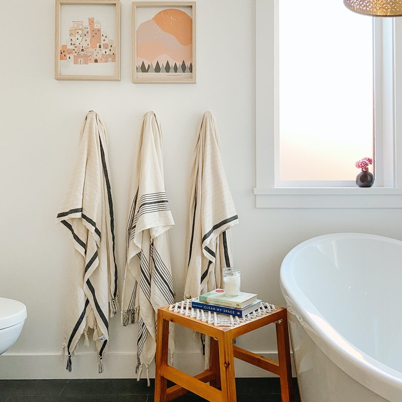 May include: Bathroom scene with three cream-colored towels with black stripes hanging on a white wall. Two framed art pieces are above the towels. A small wooden stool holds books and a candle. A white bathtub is in the background.