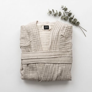 May include: A folded beige robe made of textured muslin fabric with a wrap-around tie belt and a black brand label at the inner neckline. The garment features a V-neckline and long sleeves, presented neatly with a small sprig of eucalyptus nearby.